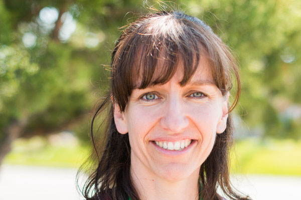 A smiling woman with dark brown hair and bangs standing outside.