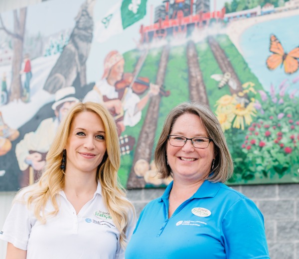 Two women stand in front of a colourful banner. One is wearing a blue Chigamik shirt..