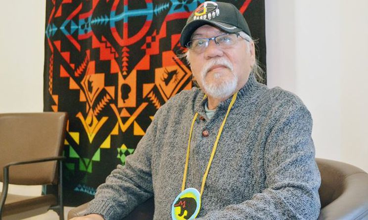 Brian George sitting in a chair with a colourful blanket on the wall behind him