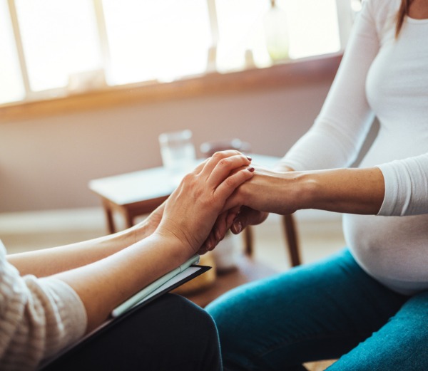 A pregnant woman sits on a chair facing another woman who is holding her hand.