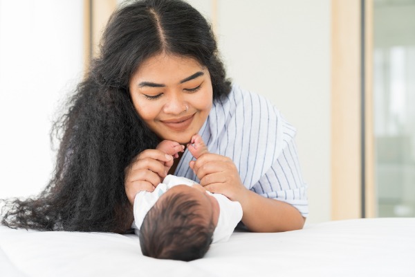 A smiling woman with long, black, curly hair leans over a very tiny baby holding its hands.