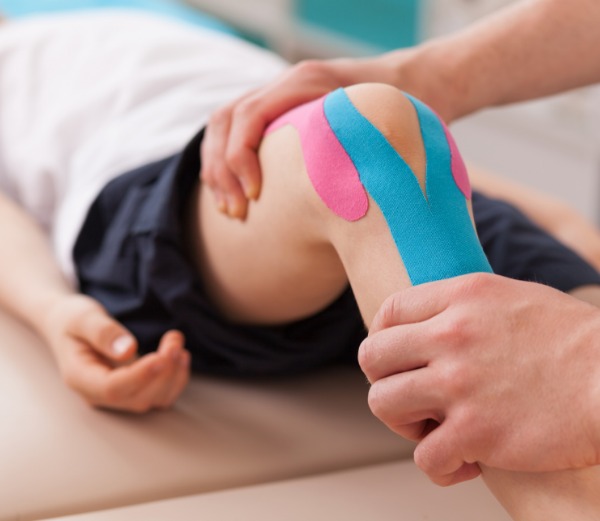 Hands working on a patients knee as they lie on a table with their knee bent with blue and pink tape wrapped on the knee.