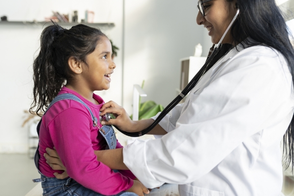 An female doctor holds a stethoscope up to the chest of a child in a pink shirt and overalls.