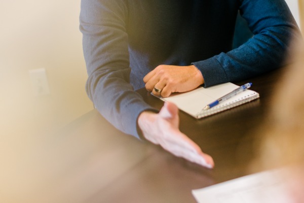 A man sits at a table with a notepad and pen. He has his hand extended ready to shake.