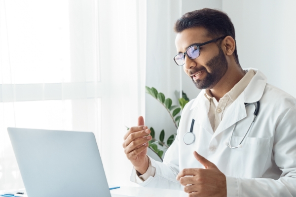 A doctor wearing glasses, a white lab coat and a stethoscope is sitting in front of a lap top speaking with someone virtually.