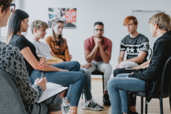 A group of six teens sit on hairs in a circle. A woman is sitting with them with a pen and clipboard.