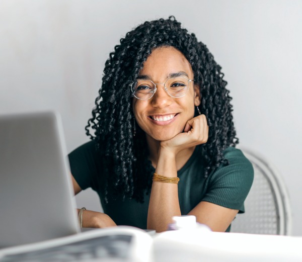 A woman with long, black, curly hair smiles for the camera while sitting at a desk in front of a computer.