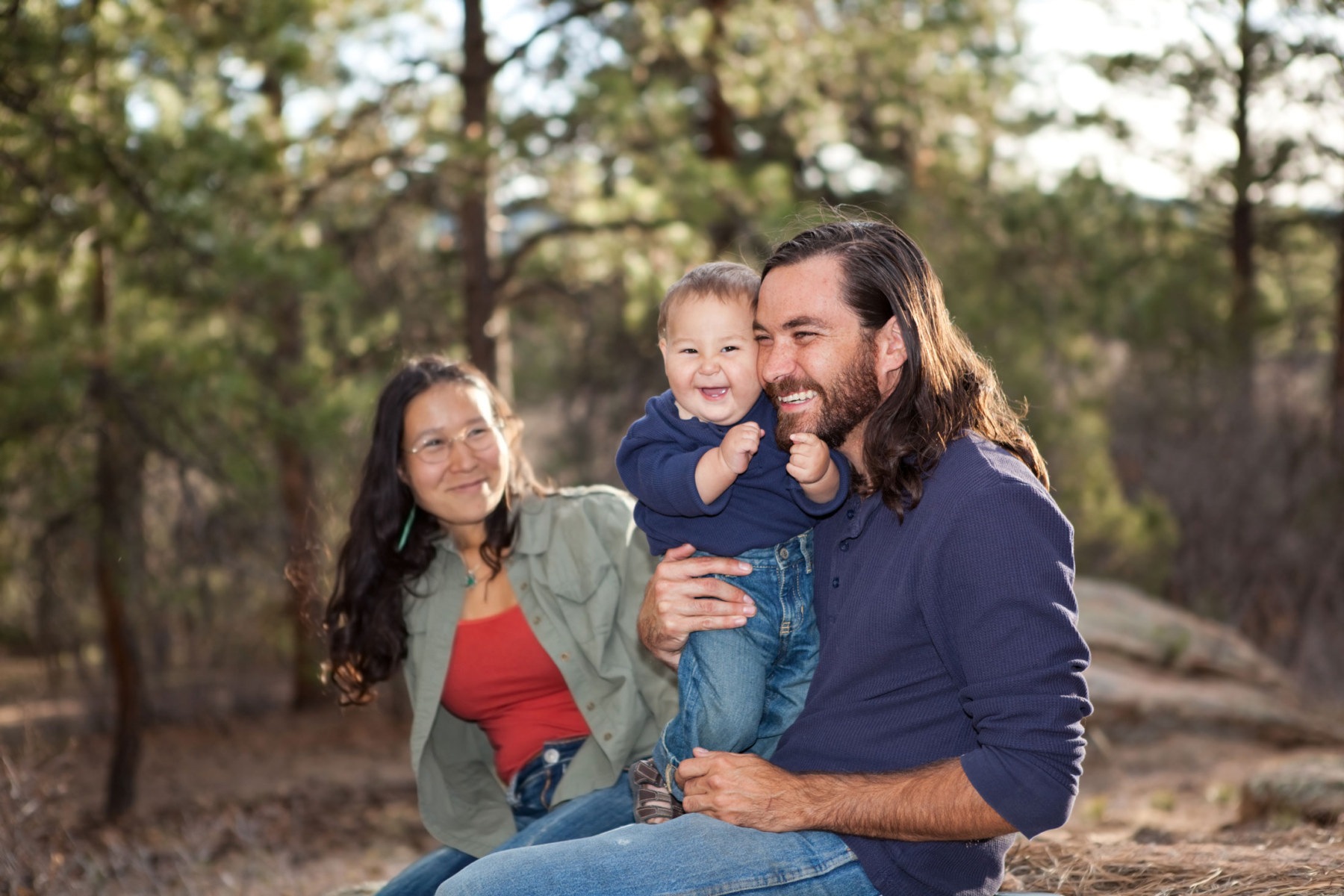 Indigenous woman and man, holding a toddler, all sitting outside in the forrest.