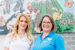 Two women stand in front of a colourful banner. One is wearing a blue Chigamik shirt..